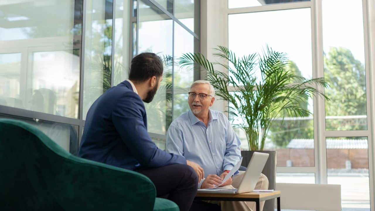 Two men are talking in a modern office, with one man holding a laptop.
