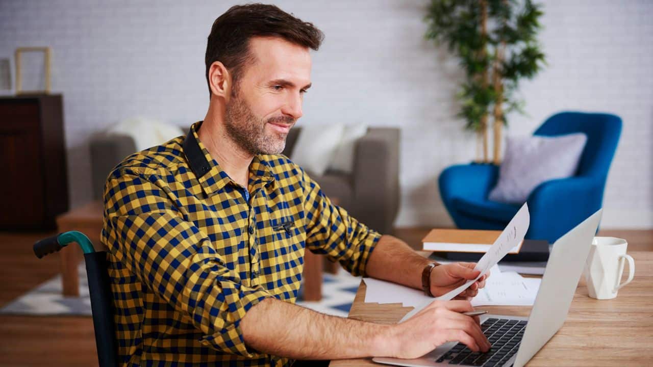 A man in a wheelchair is looking at paperwork while using his laptop at a desk.