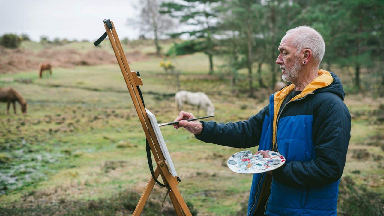 A gray-haired man in a blue jacket is painting on an easel outdoors with horses in the background.