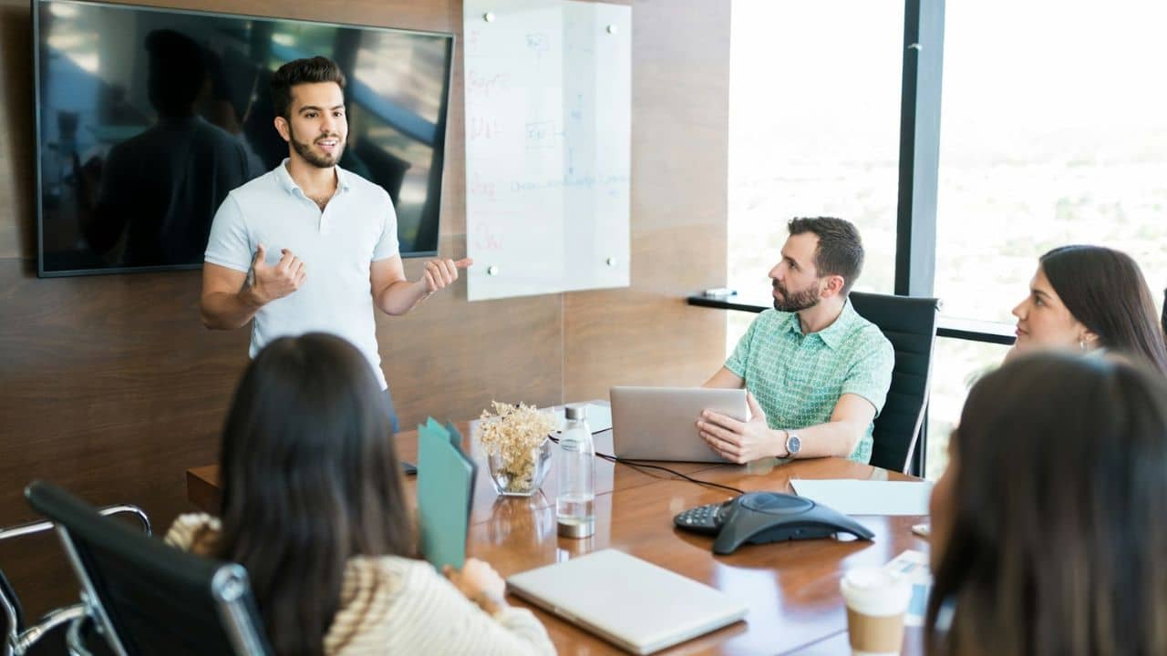 A man in a polo shirt is giving a presentation to a small group of colleagues in a conference room.