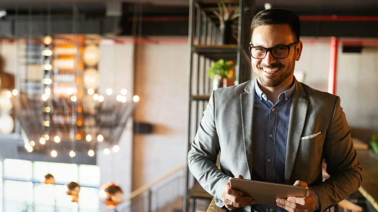 A handsome, happy businessman in a suit smiles while holding a tablet in a modern office.