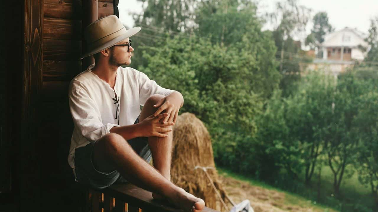 A barefoot man wearing a hat and sunglasses sits on a wooden porch, looking out at the green landscape.