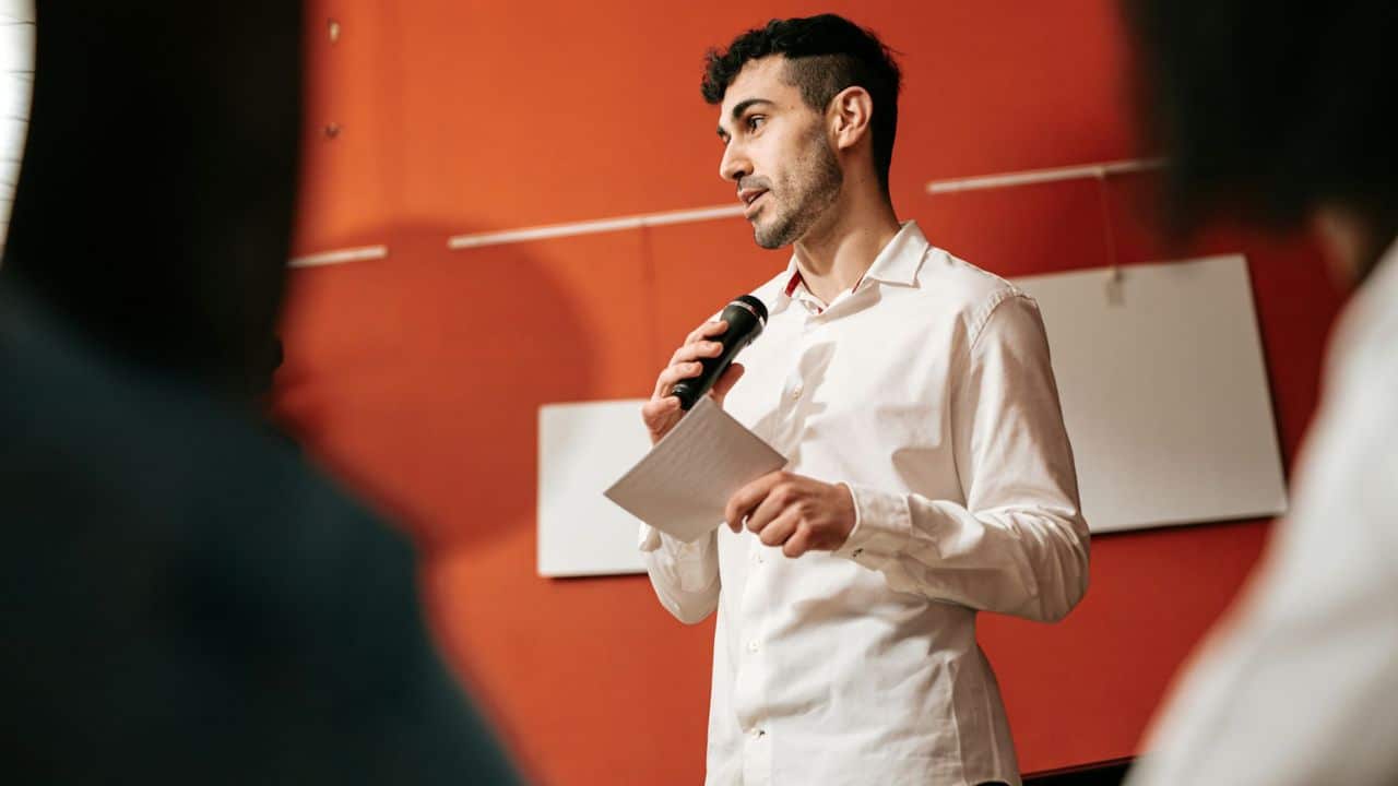 A man in a white button-down shirt is speaking into a microphone while holding a piece of paper.