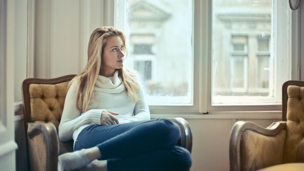 A woman in a white sweater sits by a window, looking thoughtfully to the side.