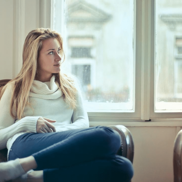 A woman in a white sweater sits by a window, looking thoughtfully to the side.