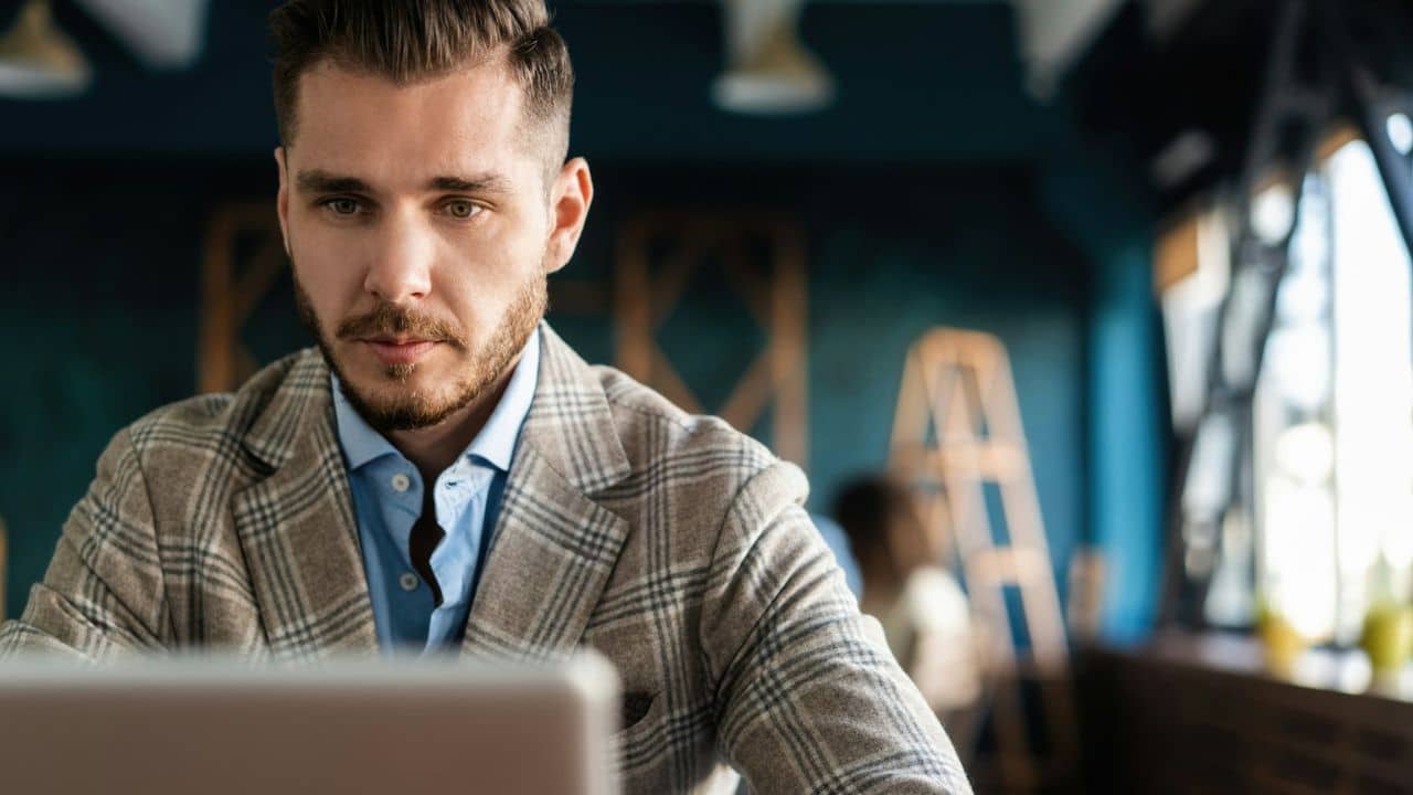 A handsome man with a plaid jacket is sitting at a desk, looking down at his laptop.