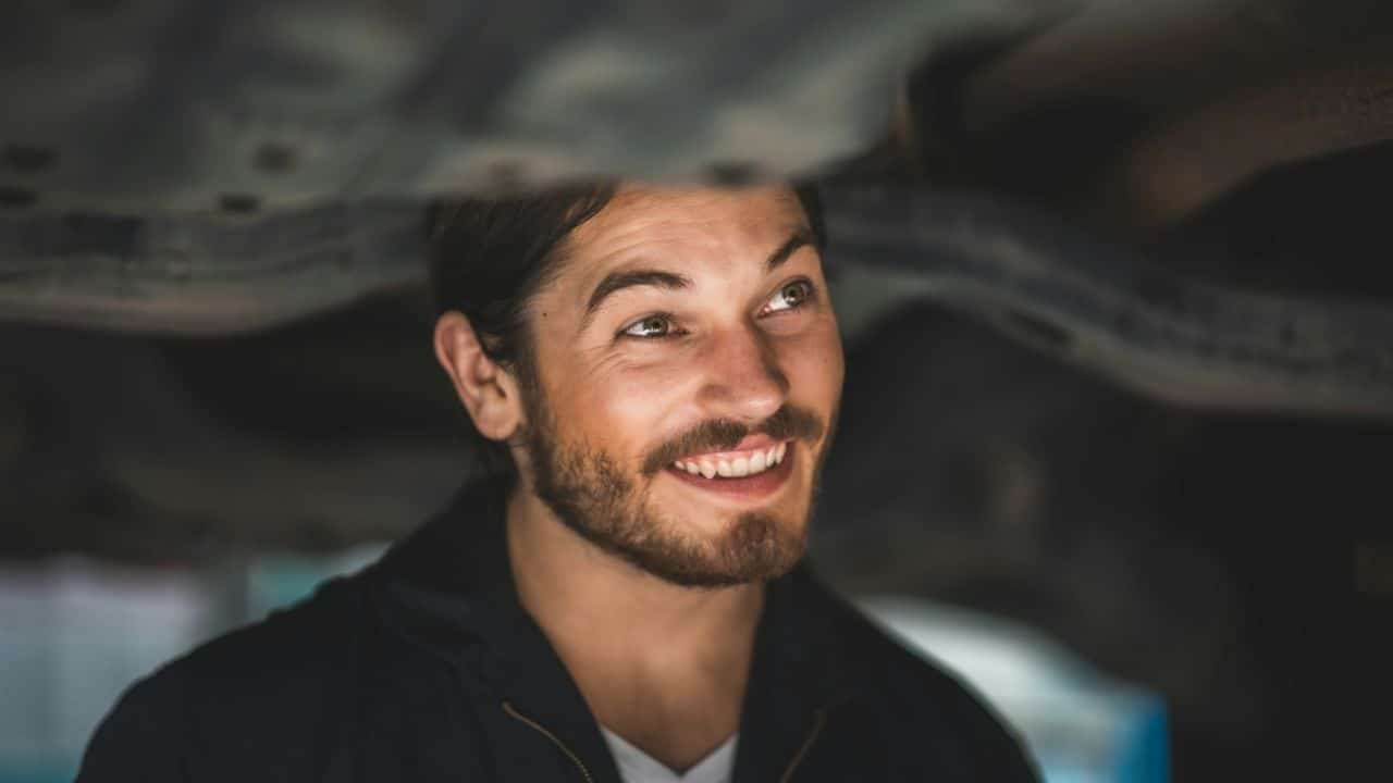 A handsome, happy mechanic with a beard is smiling while working underneath a car.