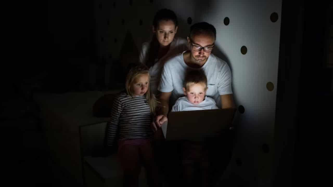 Man working on laptop late at night while family is visible nearby.