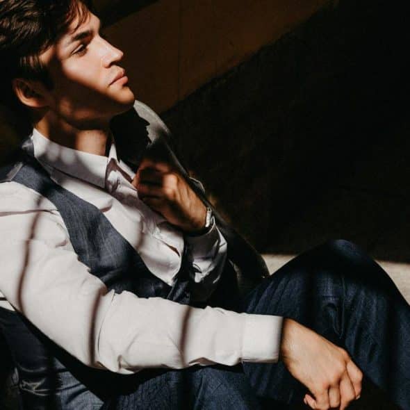 A young man in a suit sits against a wall with sunlight casting shadows across his face.