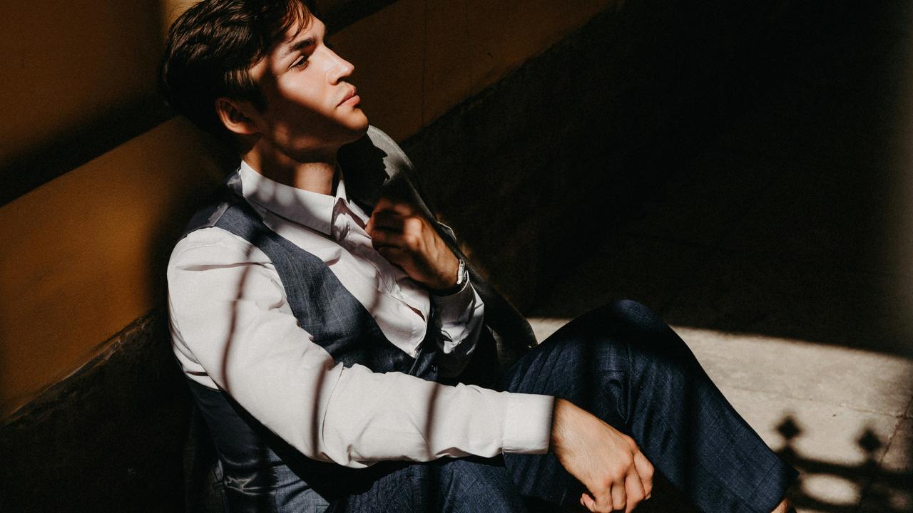 A young man in a suit sits against a wall with sunlight casting shadows across his face.