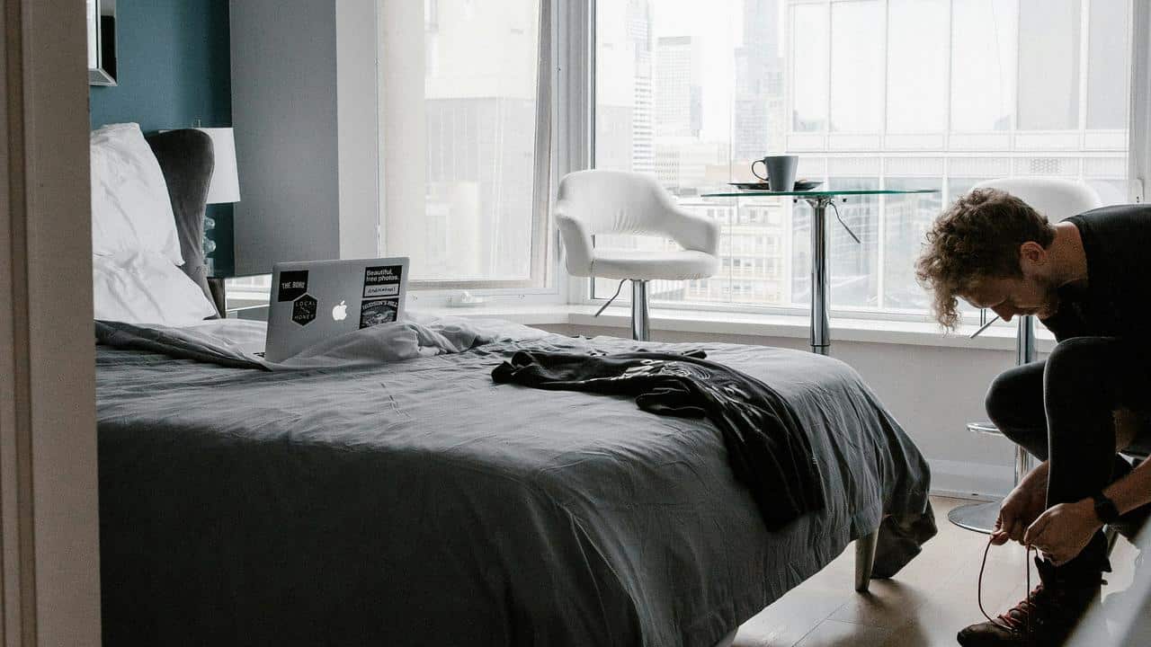 A man ties his shoes in a bedroom with a laptop on the bed.