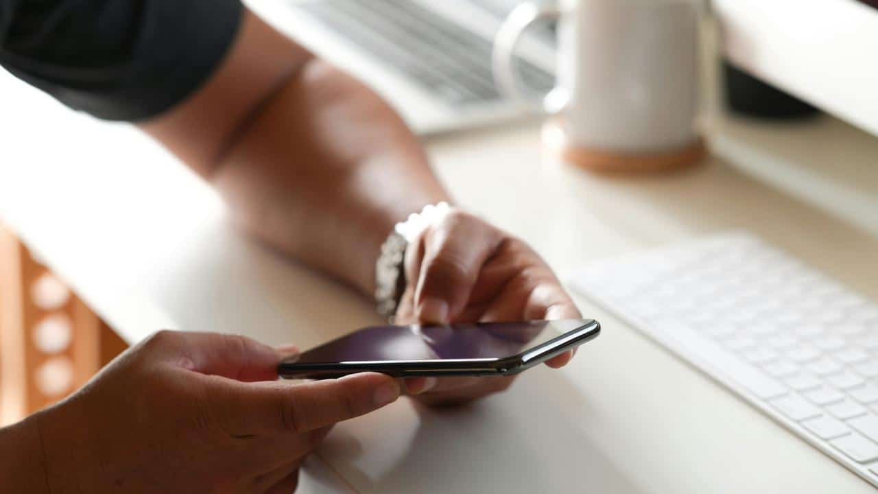 A person holds a smartphone while sitting at a desk with a keyboard and mug.