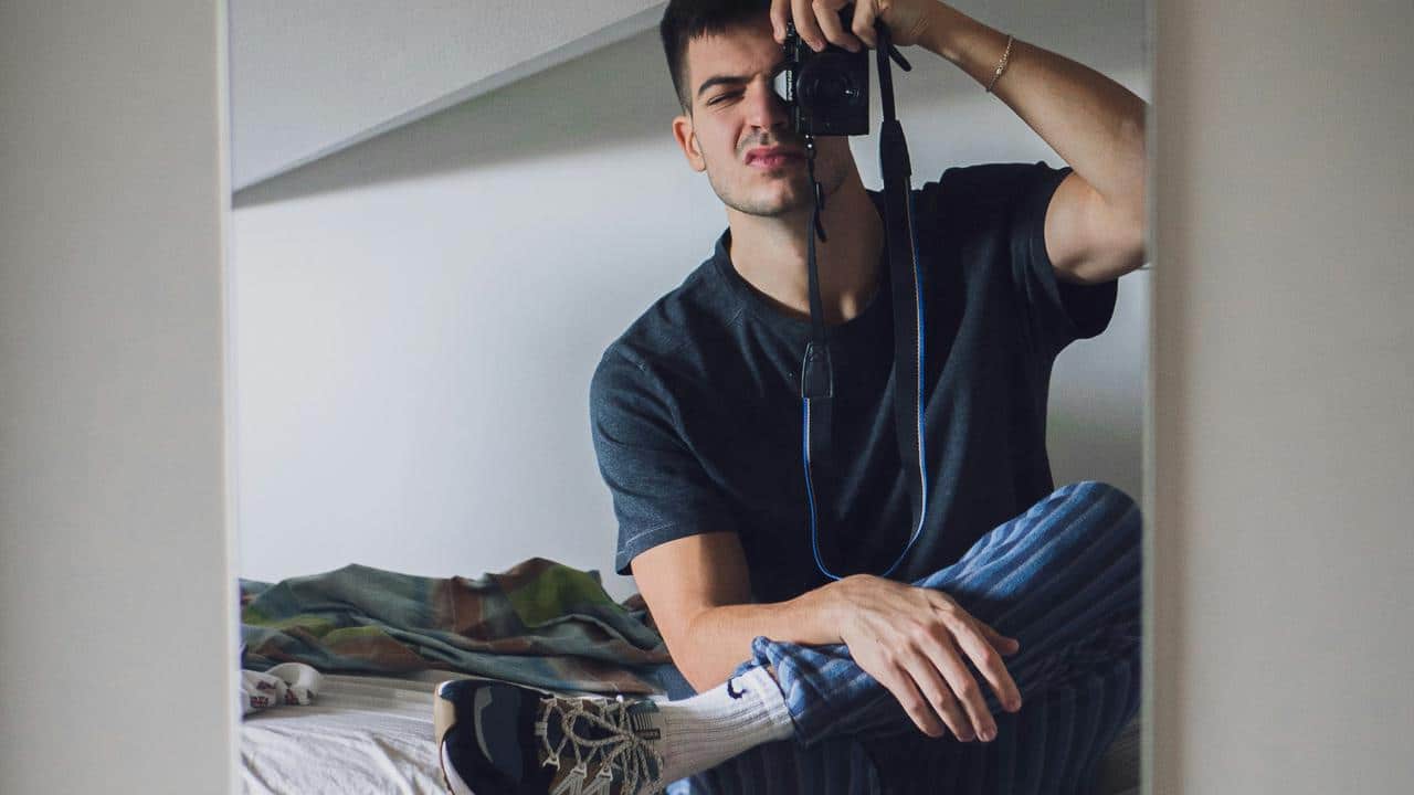 A man sits on a bed taking a mirror selfie with a camera.