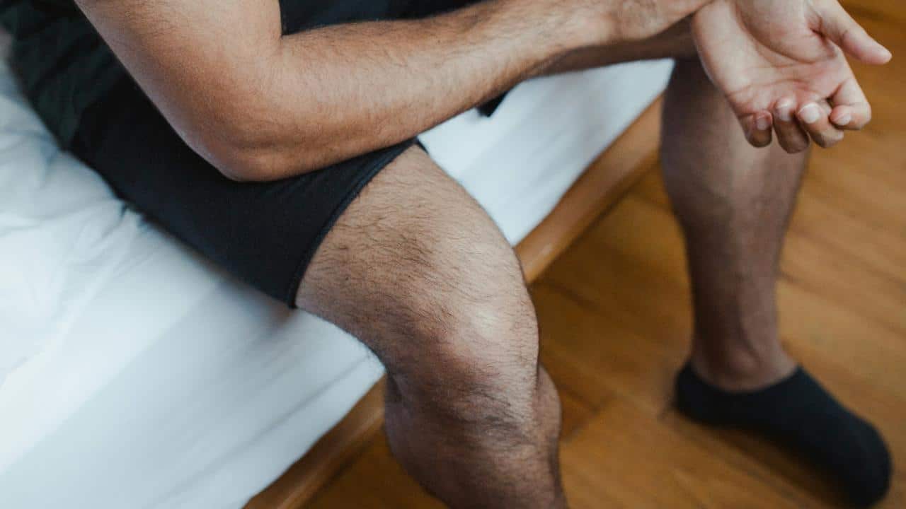 A person in black shorts and socks sits on a bed with hands resting on their lap.