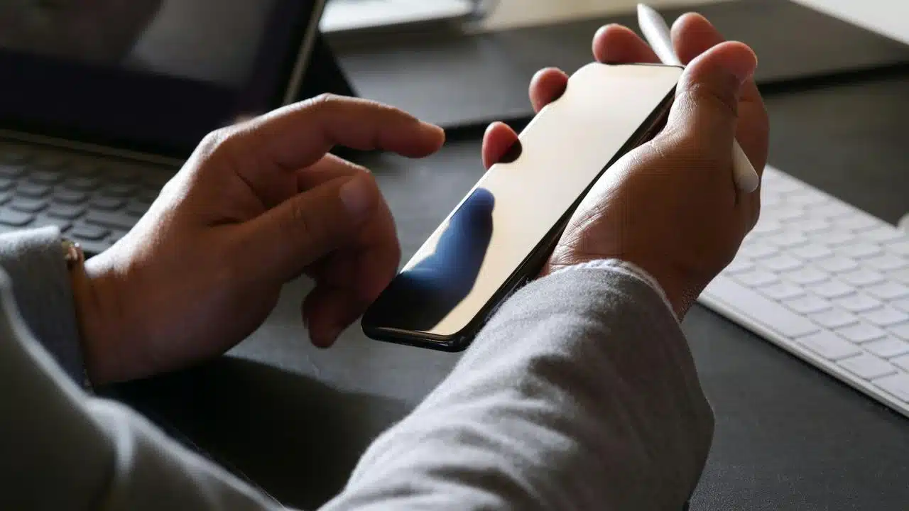 A person holding a smartphone and stylus while using a laptop and keyboard.