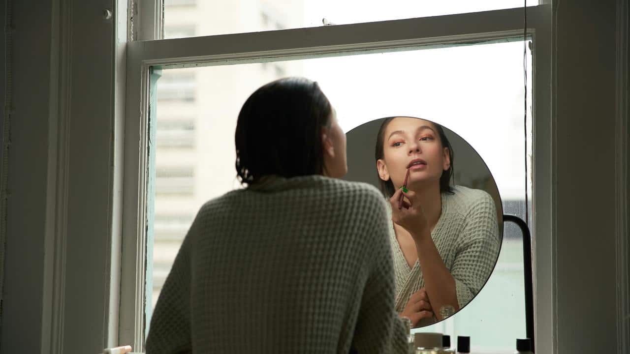 A woman applying lipstick while looking into a round mirror.