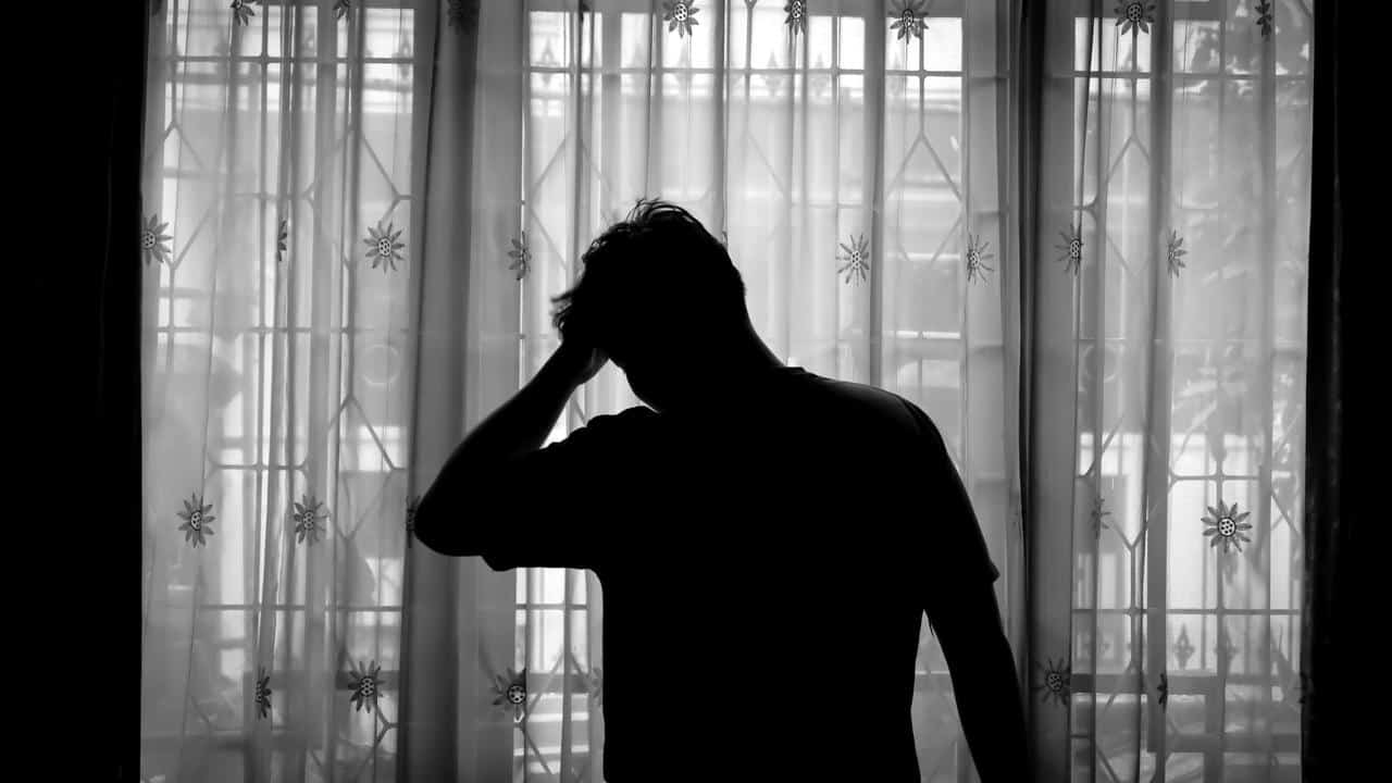 A silhouette of a person standing by a window with one hand on their head.