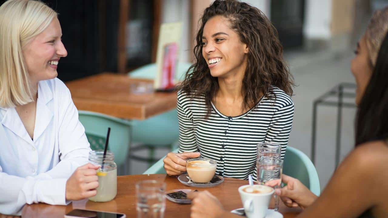A group of women smiling and chatting over coffee at an outdoor café.