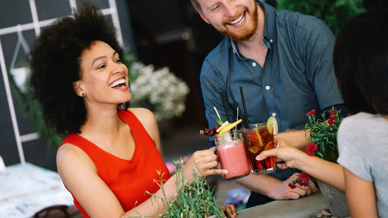 A group of friends laughing and clinking glasses with colorful drinks outdoors.