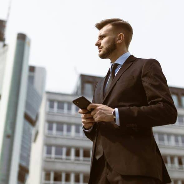 A man in a suit holding a phone in front of tall office buildings.