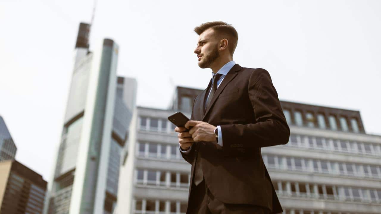 A man in a suit holding a phone in front of tall office buildings.