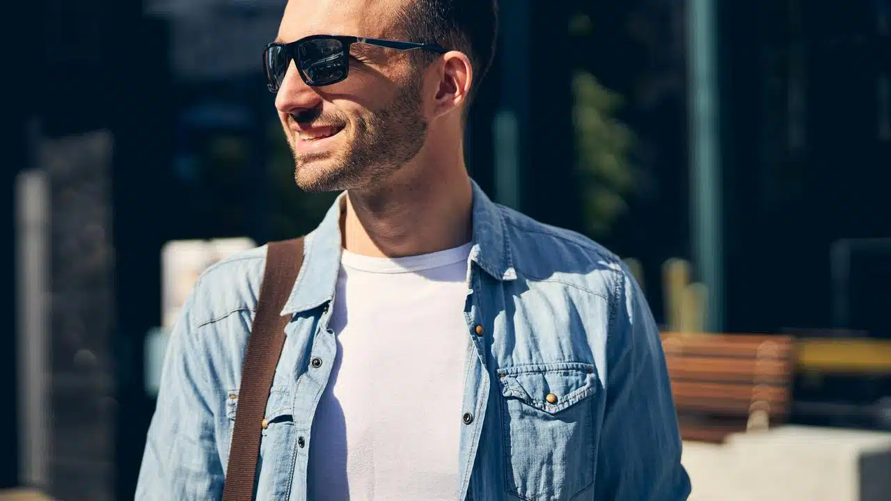 A man in sunglasses wearing a denim shirt and white t-shirt outdoors.