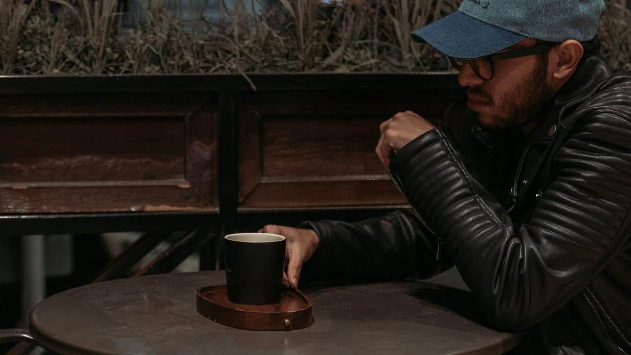 A man in a leather jacket and cap sitting at a table with a cup of coffee.
