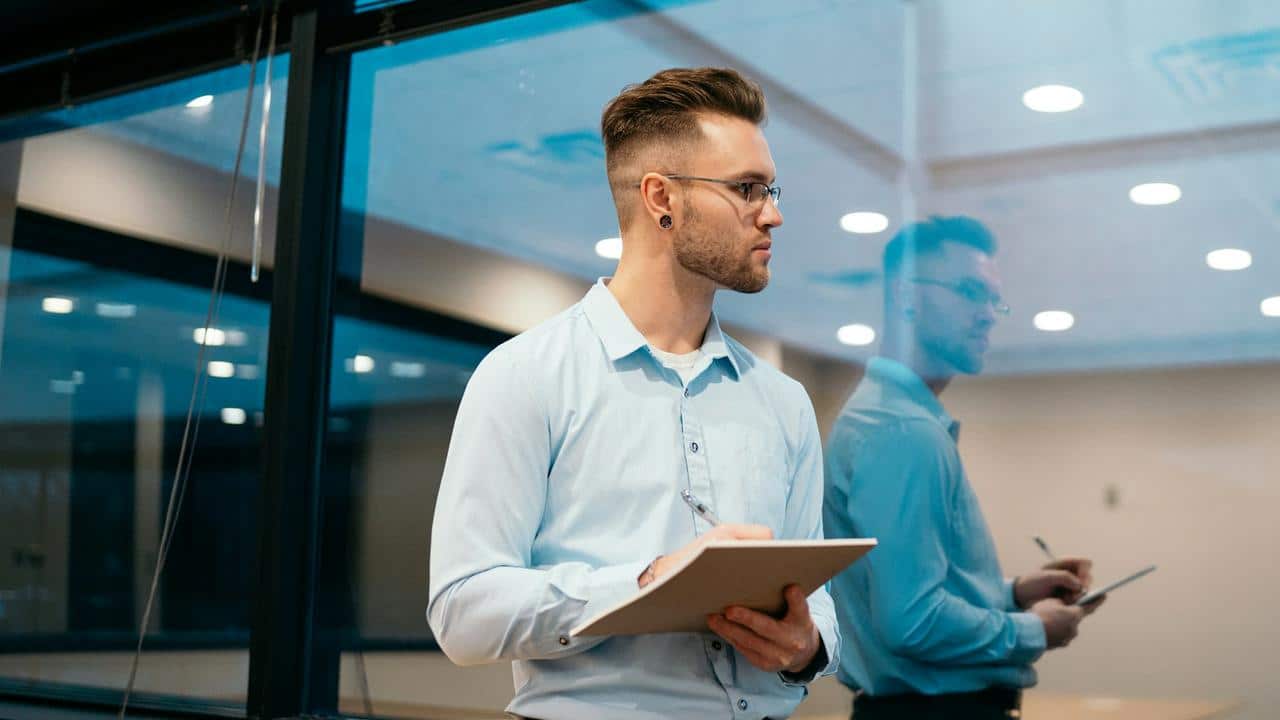 A man in glasses holding a tablet while standing in an office.