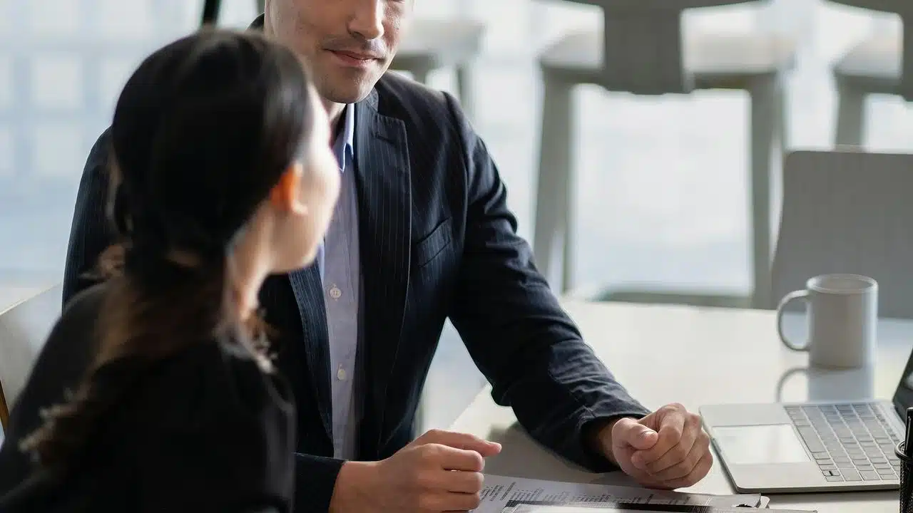 A businessman talking with a colleague at a desk with documents and a laptop.