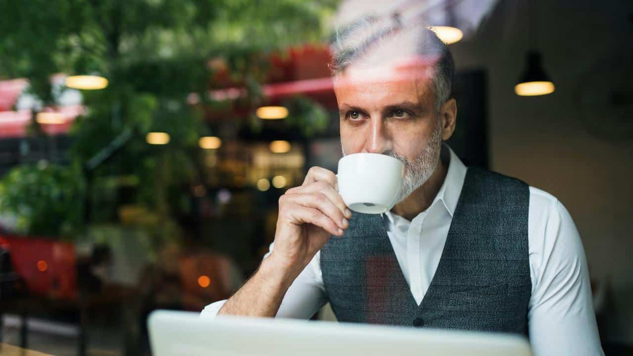 A man with gray hair and beard drinking coffee while sitting at a cafe table.