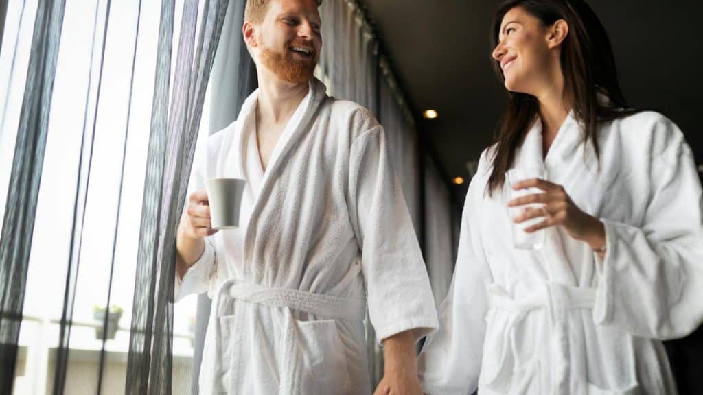 A couple in white bathrobes smiling and holding drinks indoors.