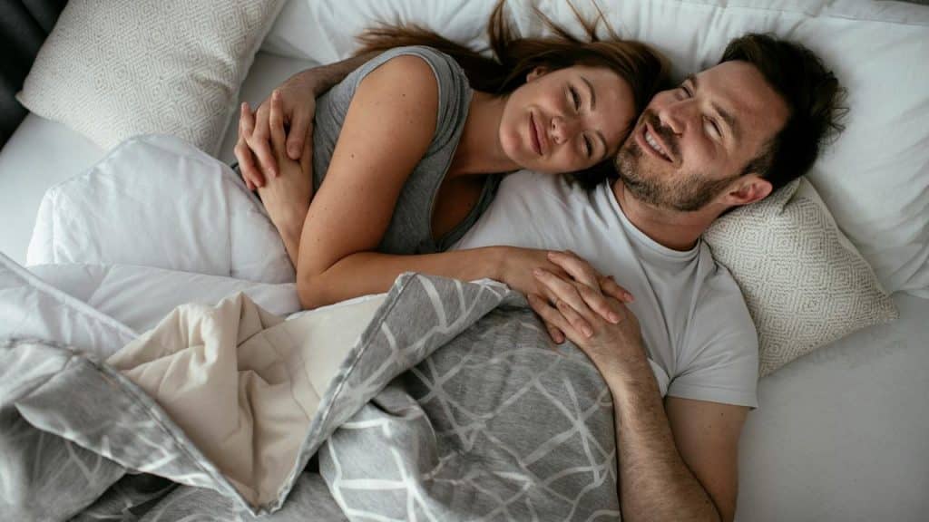 A couple cuddling in bed while smiling and holding hands.