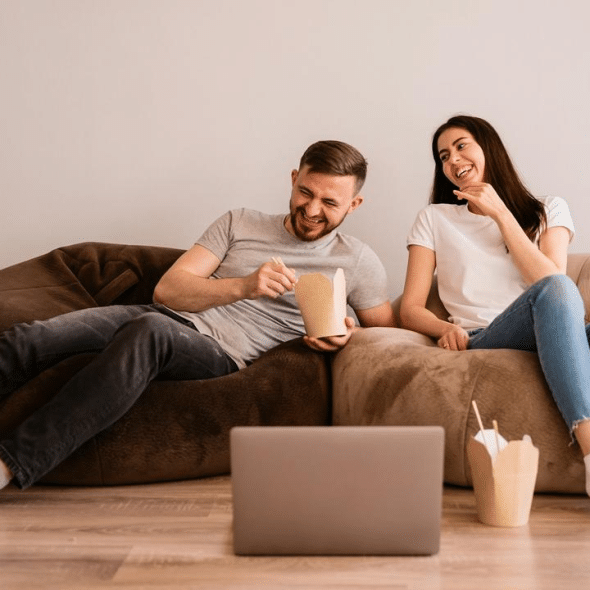 A couple sitting on bean bags eating takeout while watching a laptop.
