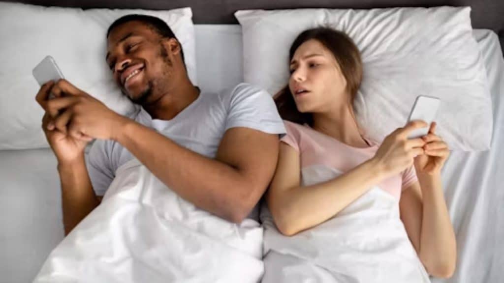 A couple lying in bed facing away from each other and busy with their screens