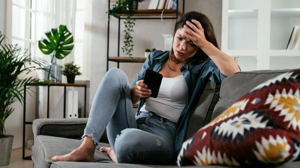 A woman sitting on a couch looking stressed while holding a smartphone.