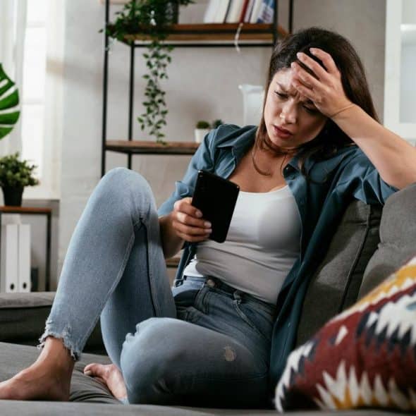 A woman sitting on a couch looking stressed while holding a smartphone.
