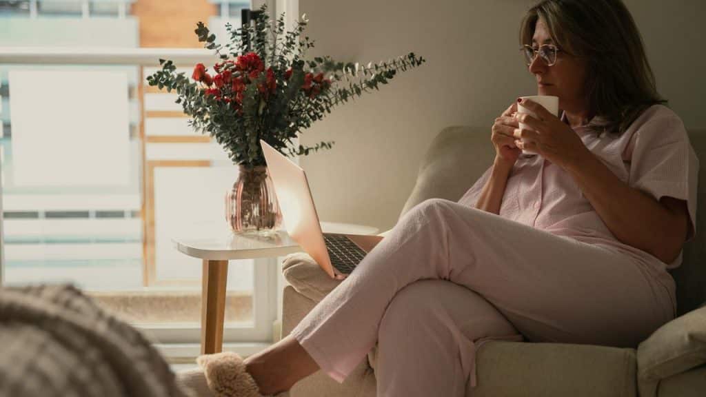 A woman sitting on a couch in pajamas holding a cup and looking at a laptop.