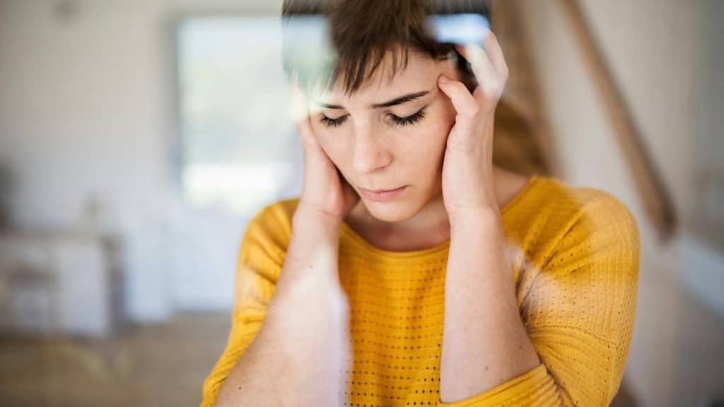 A woman in a yellow sweater holding her head with both hands and looking down.