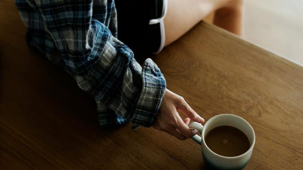 A person in a plaid shirt holding a cup of coffee on a wooden table.