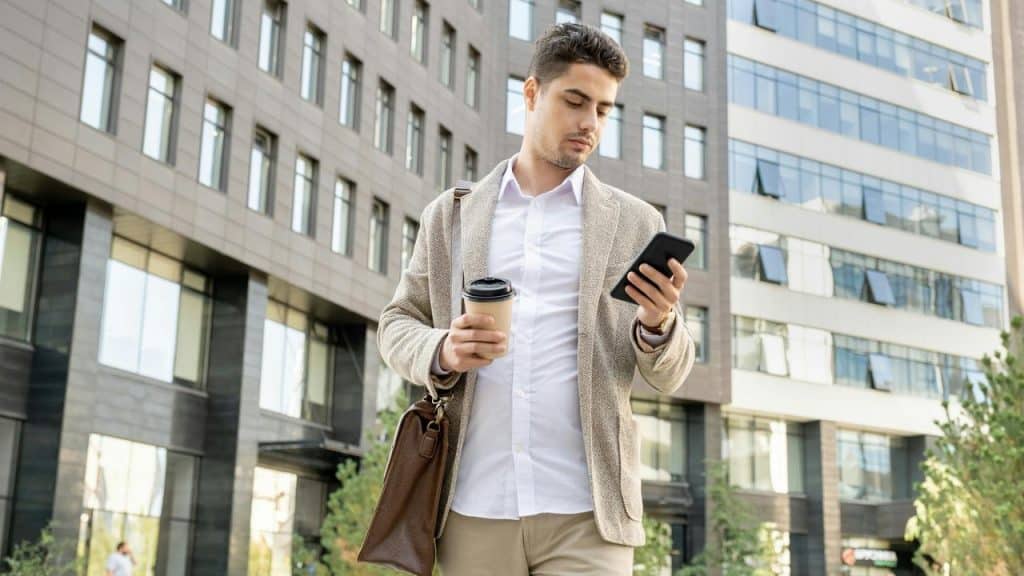 A man walking outside holding coffee and looking at his phone.