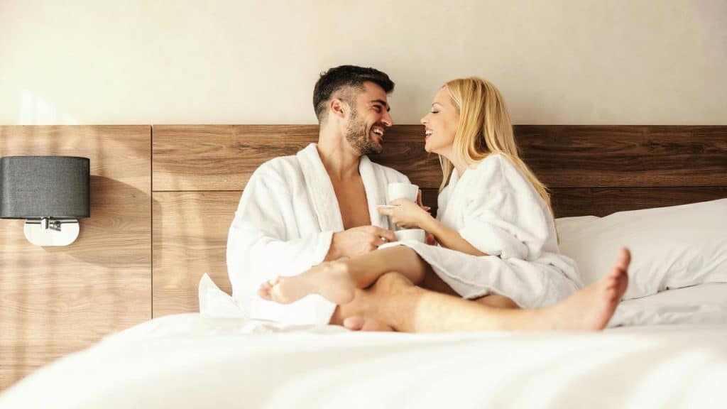 A couple in robes smiling and drinking coffee in bed.