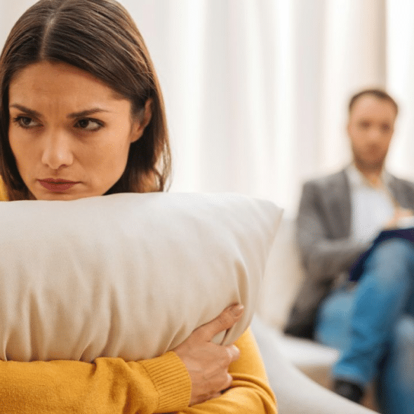 A concerned woman in a yellow sweater is holding a pillow and looking to the side, with a man blurred in the background.