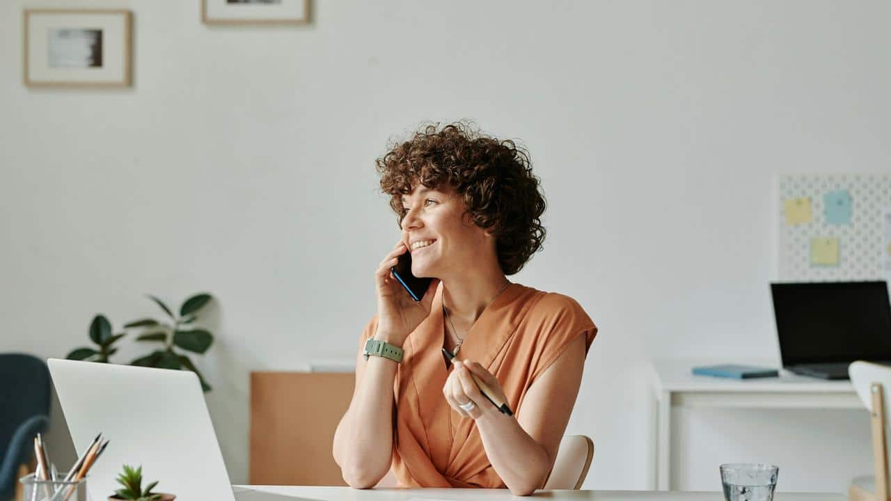A woman smiling while talking on the phone at her desk.