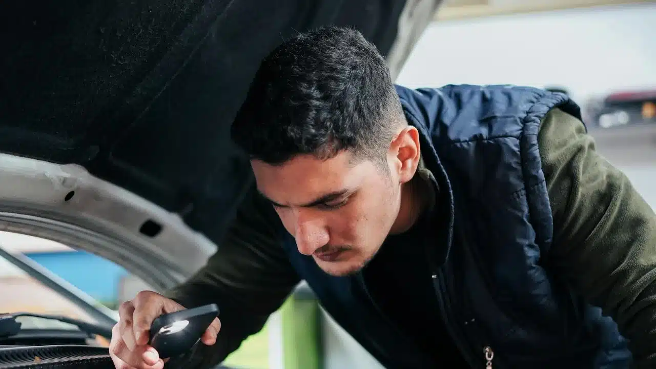A man inspecting a car engine with a flashlight on his phone.