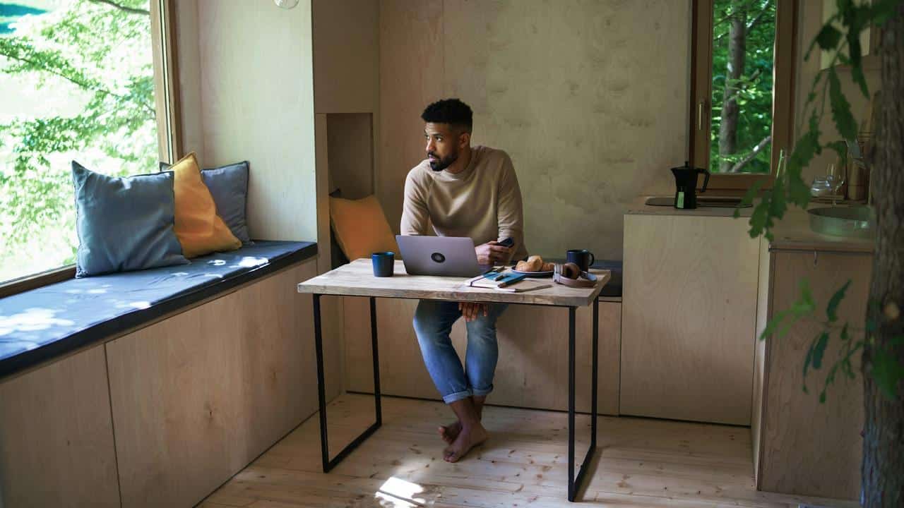 A man working on a laptop at a wooden desk in a cozy room with large windows.