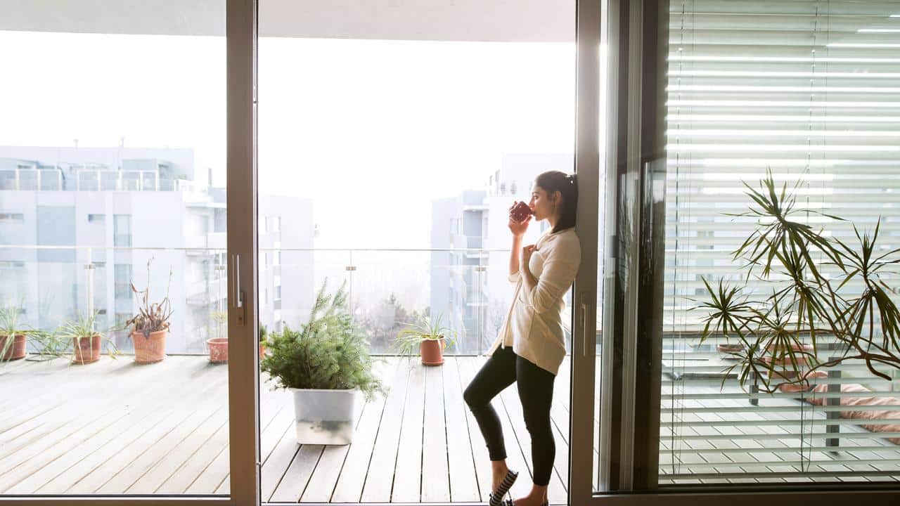 A woman drinking from a cup while standing by a glass door on a balcony.