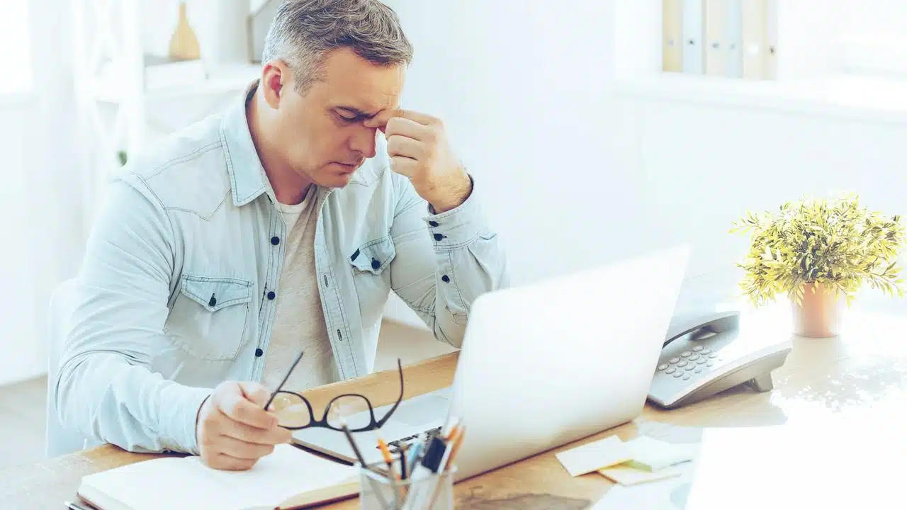 A man sitting at a desk holding his glasses and rubbing his forehead in frustration.