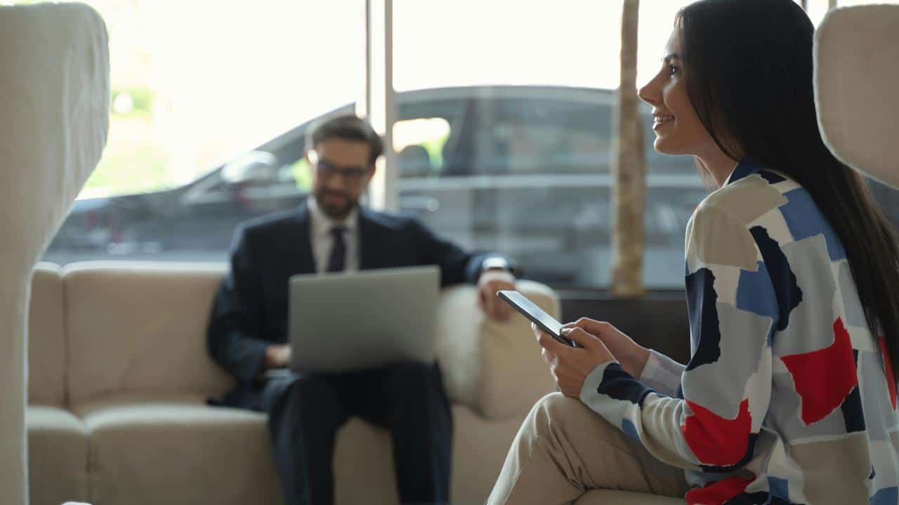 A woman holding a tablet and smiling while a man works on a laptop in the background.