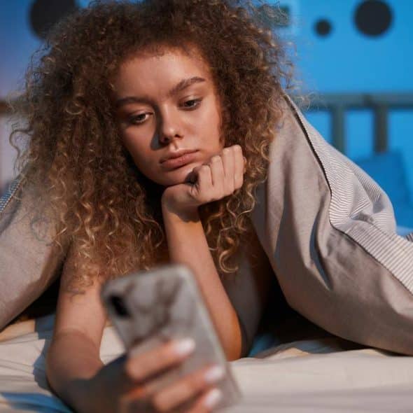 A young woman lying in bed looks at her phone with a thoughtful expression.
