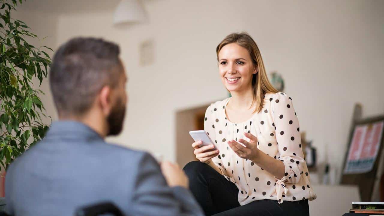 A smiling woman holding a phone talks to a man in an indoor setting.